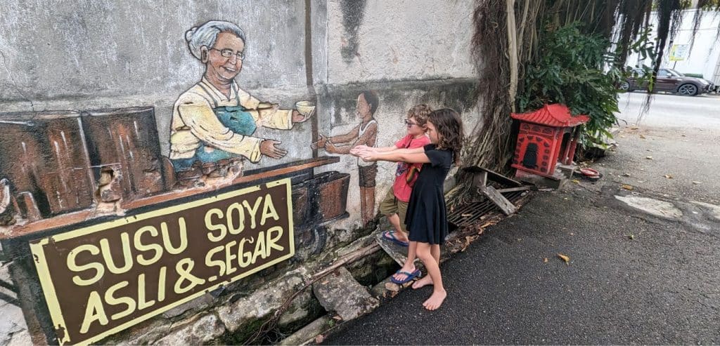 Two kids pretending to accept treats from a street painting of a vendor in Georgetown, Penang.